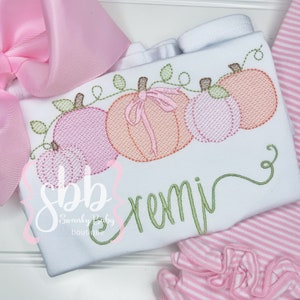 May include: White baby shirt with embroidered pumpkins in shades of pink and orange, with green vines and leaves. The name "remi" is embroidered in green script. Pink striped pants and a pink bow are also visible.