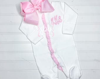 May include: White baby footed sleeper with pink ruffle trim and embroidered initials. A large pink bow is attached at the top. The sleeper has long sleeves and snaps down the front.