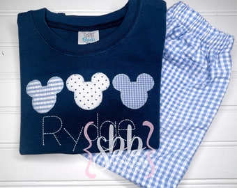 Puede incluir: Una camiseta azul marino con tres orejas de Mickey Mouse bordadas en diferentes patrones. La camiseta tiene el nombre "Ryder" bordado debajo de las orejas. La camiseta se combina con pantalones de cuadros azules y blancos.