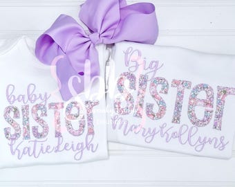 Puede incluir: Dos camisetas blancas con letras de estampado floral que dicen baby SISTER Katie Leigh y Big SISTER Mary Kollyns en letra cursiva morada clara. Un lazo grande de color morado claro está colocado sobre las camisetas en un fondo blanco.