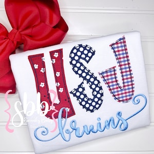 May include: White t-shirt with the letters "USJ" embroidered in red, white, and blue fabric. The letters are decorated with polka dots and gingham patterns. The text "twins" is embroidered in blue below the letters.