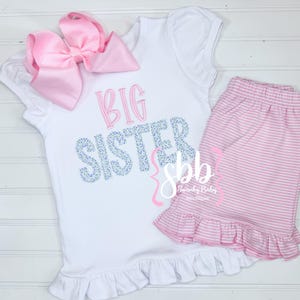 May include: White shirt with a pink bow and the text "BIG SISTER" in blue and white. The shirt has a ruffle at the bottom. Pink and white striped shorts with a ruffle at the bottom.