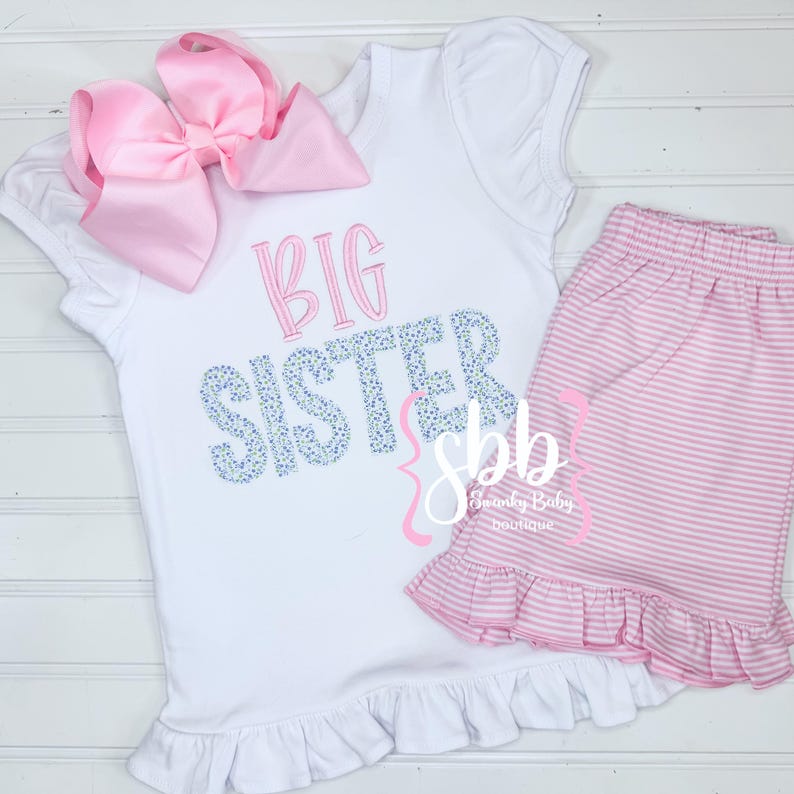 May include: White shirt with a pink bow and the text "BIG SISTER" in blue and white. The shirt has a ruffle at the bottom. Pink and white striped shorts with a ruffle at the bottom.