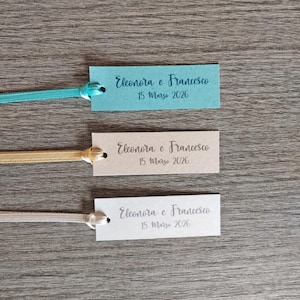 Può includere: Tre etichette rettangolari con la scritta "Eleonora e Francesco 15 Marzo 2026" in elegante corsivo. Ogni etichetta è attaccata a un nastro. Le etichette sono azzurro chiaro, beige e bianche.
