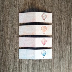 Può includere: Quattro etichette di carta piegate con illustrazioni di mongolfiere ad acquerello. Ogni etichetta presenta un diverso design di palloncino nei toni del rosso, blu e pesca. Le etichette sono disposte su una superficie di legno.