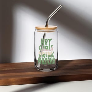 Puede incluir: Una lata de vidrio transparente con tapa de bambú y una pajita de metal. La lata tiene un gráfico verde y rosa que dice "Hot Girls Drink Matcha".