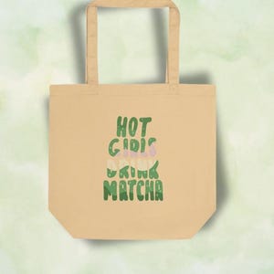 May include: A beige tote bag with two shoulder straps featuring the green text HOT GIRLS DRINK MATCHA printed in a wavy, stacked font. The design includes small star accents within the letters, creating a layered, retro-inspired graphic.
