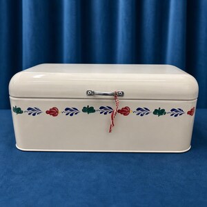 Op de afbeelding: Een crèmekleurige rechthoekige broodtrommel van metaal met afgeronde hoeken en een zilveren handvat. De voorzijde is voorzien van een horizontale decoratieve rand met afwisselend rode, blauwe en groene bladachtige patronen.
