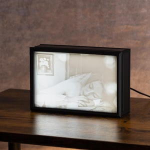 May include: A black framed light box with a picture of a person sleeping in a bed. The picture is in black and white and the light box is illuminated.