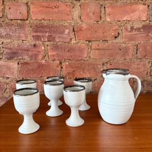 May include: A white ceramic jug and six matching goblets with a dark brown rim. The jug has a handle and a spout. The goblets have a pedestal base. The set is on a wooden surface against a brick wall.