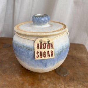 May include: A round ceramic jar with a lid, featuring a cream and blue ombre glaze. A light-coloured string is wrapped around the neck, holding a rectangular ceramic tag with the words BROWN SUGAR embossed in dark red lettering on the front.