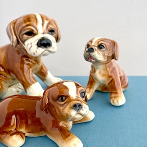 May include: Three ceramic dog figurines in brown and white. One is lying down, another is sitting, and the third is standing. The figurines have glossy finishes and are set on a blue surface. The dogs are in various poses.