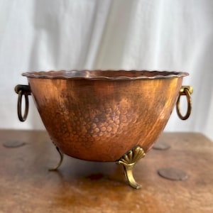 May include: A round copper bowl featuring a hammered texture, a scalloped rim, and two side ring handles. The bowl is supported by three decorative brass feet shaped like shells, providing a stable base for the metallic vessel.