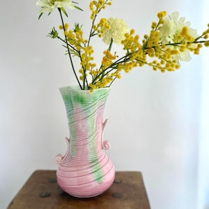 May include: A pink and green ceramic vase with a wavy design, holding yellow and white flowers. The vase has a bulbous base and two curved handles. The flowers add a pop of color to the arrangement.