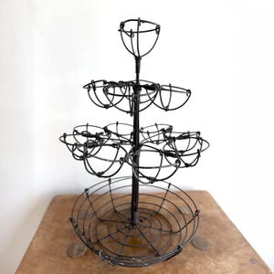 May include: A tiered black wire display stand featuring a large circular base basket at the bottom, a middle section with four smaller wire cup holders, a second tier with two wire cup holders, and a single wire cup holder at the very top.