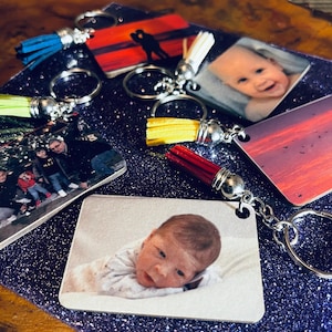 Op de afbeelding: Sleutelhangers met gepersonaliseerde foto-afdrukken. De sleutelhangers tonen verschillende afbeeldingen, waaronder een baby, een silhouet van een stel tegen een zonsondergang en een gezin. Elke sleutelhanger heeft een metalen ring en een kwast in verschillende kleuren.