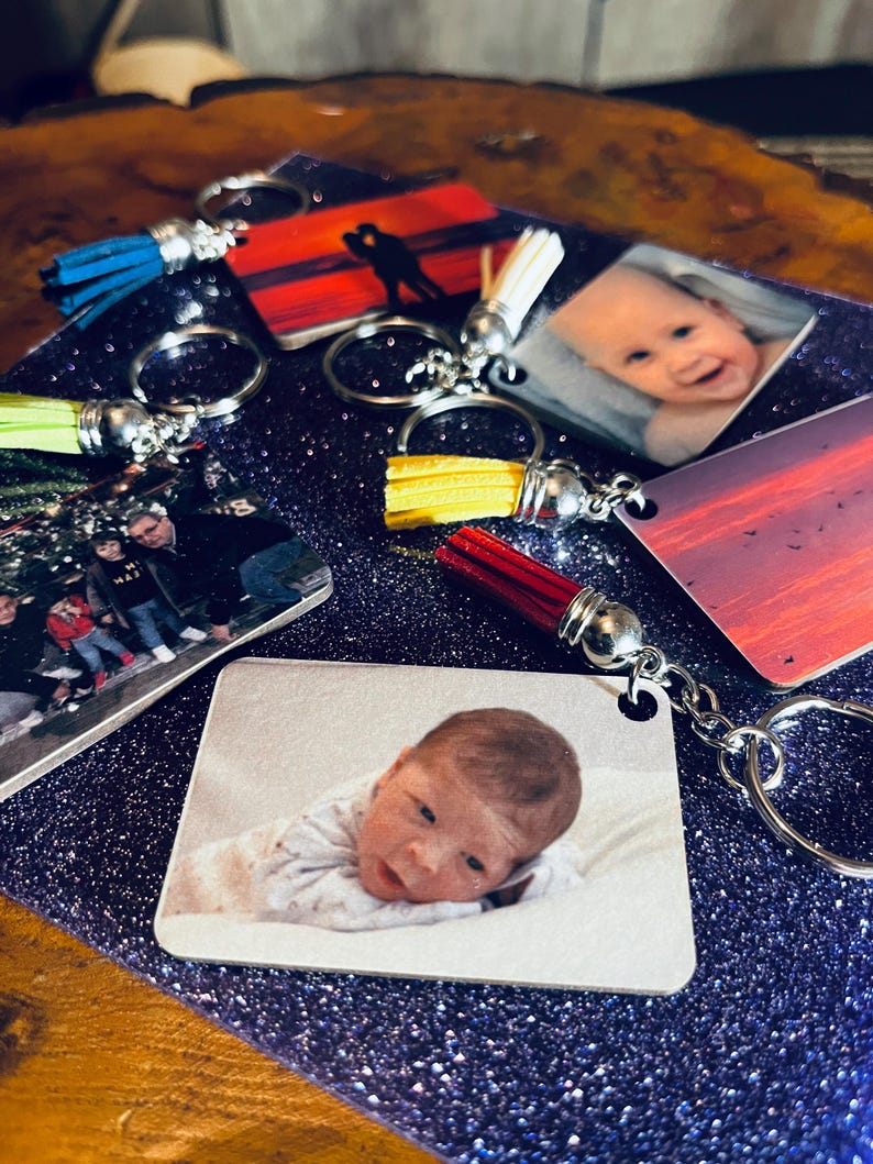 Op de afbeelding: Sleutelhangers met gepersonaliseerde foto-afdrukken. De sleutelhangers tonen verschillende afbeeldingen, waaronder een baby, een silhouet van een stel tegen een zonsondergang en een gezin. Elke sleutelhanger heeft een metalen ring en een kwast in verschillende kleuren.