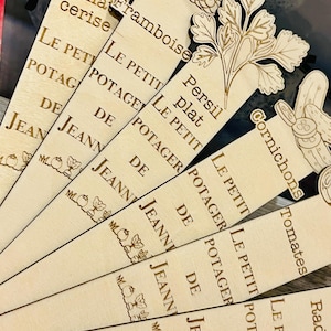Puede incluir: Marcadores de madera con ilustraciones botánicas grabadas y texto. Los marcadores presentan diseños de tomates, frambuesas, perejil, pepinos y rábanos. El texto incluye las palabras "LE PETIT POTAGER DE JEANNE!"