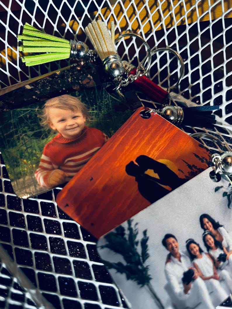 Op de afbeelding: Een verzameling gepersonaliseerde sleutelhangers met kwastjes in verschillende kleuren, waaronder groen, rood en beige. De sleutelhangers hebben afgedrukte foto's van een kind, een stel bij zonsondergang en een gezin. De sleutelhangers zijn bevestigd aan zilveren sleutelringen.
