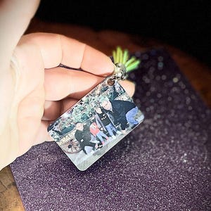 Op de afbeelding: Een rechthoekige sleutelhanger met een familiefoto, een zilverkleurige metalen lus en een groene accent. De sleutelhanger wordt tegen een paars, glinsterend oppervlak gehouden.
