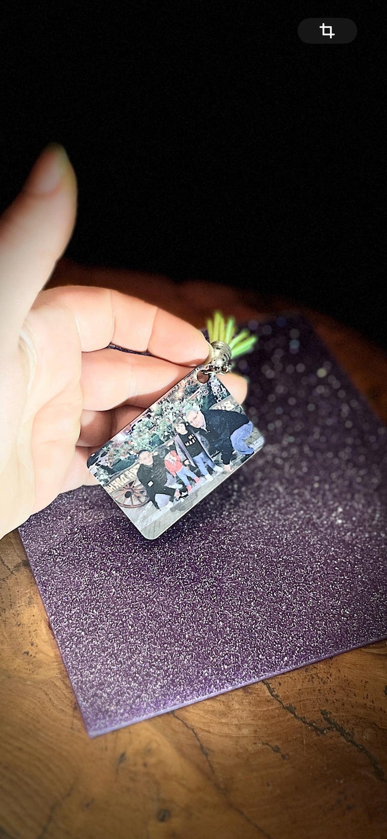 Op de afbeelding: Een rechthoekige sleutelhanger met een familiefoto, een zilverkleurige metalen lus en een groene accent. De sleutelhanger wordt tegen een paars, glinsterend oppervlak gehouden.