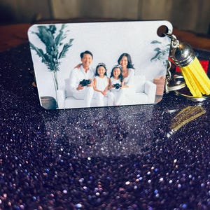 Op de afbeelding: Een vierkante sleutelhanger met een familiefoto. De afbeelding toont een gezin van vier in witte kleding, met camera's. De sleutelhanger heeft een gele kwast en zilveren hardware.