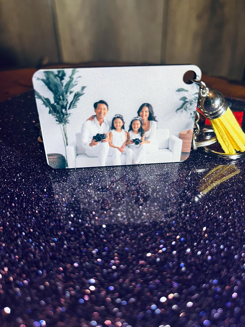Op de afbeelding: Een vierkante sleutelhanger met een familiefoto. De afbeelding toont een gezin van vier in witte kleding, met camera's. De sleutelhanger heeft een gele kwast en zilveren hardware.