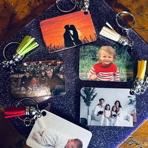 Op de afbeelding: Rechthoekige sleutelhangers met diverse foto's, waaronder een stel dat kust bij zonsondergang, een baby, een gezin en een kind in een rood-wit gestreept shirt. Elke sleutelhanger heeft een zilveren ring en een kwast in verschillende kleuren.