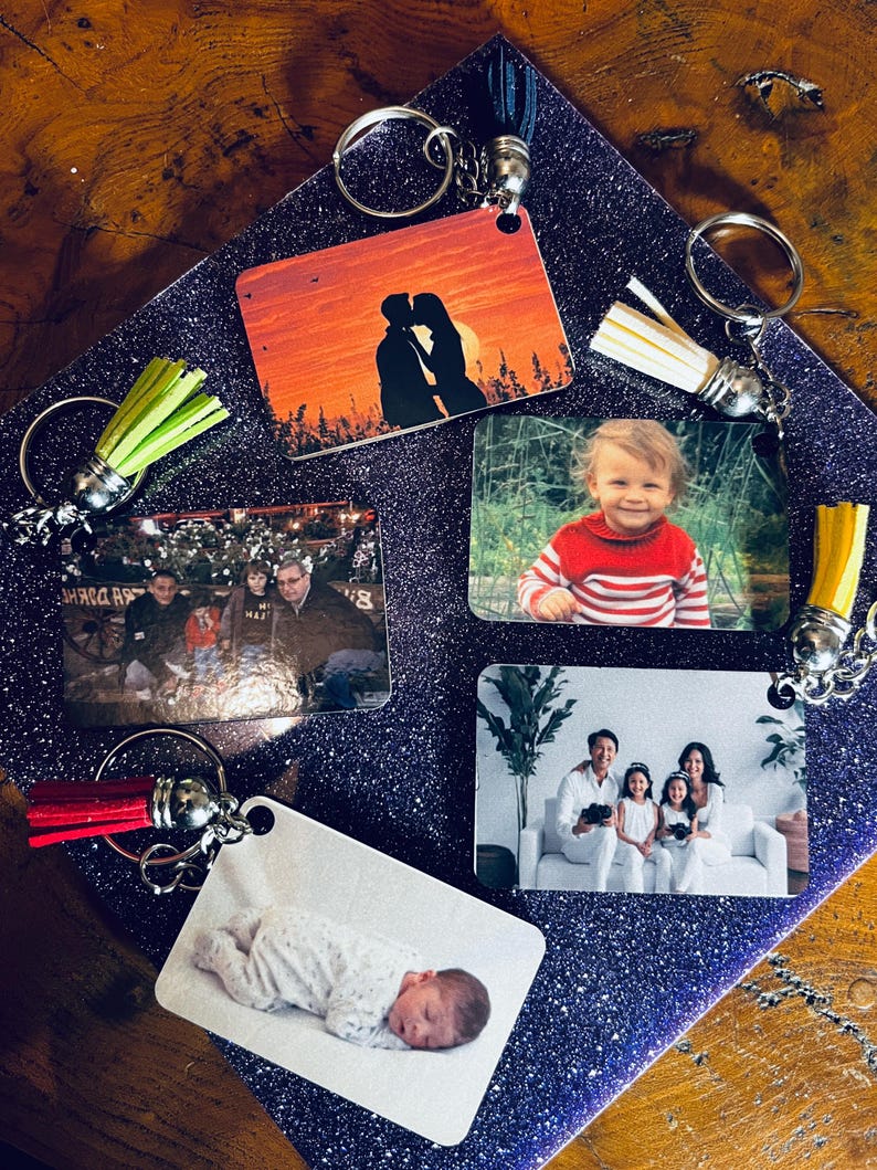 Op de afbeelding: Rechthoekige sleutelhangers met diverse foto's, waaronder een stel dat kust bij zonsondergang, een baby, een gezin en een kind in een rood-wit gestreept shirt. Elke sleutelhanger heeft een zilveren ring en een kwast in verschillende kleuren.