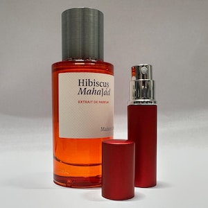 May include: A perfume bottle and a smaller atomizer. The perfume bottle is filled with a reddish-orange liquid and has a silver cap. The label reads "Hibiscus Mahajád, Extrait de Parfum". The atomizer is red with a silver top, and a red cap is next to it.