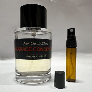 May include: A clear glass perfume bottle with a black cap, a smaller amber-coloured spray bottle, and a detached black cap. The perfume bottle has a black label with white text that reads "BIGARADE CONCENTREE" and "Jean-Claude Ellena".