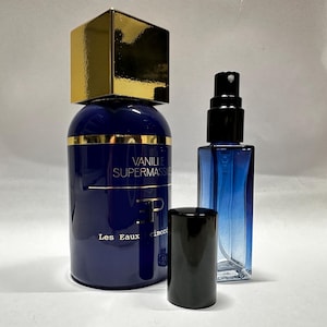 May include: A dark blue perfume bottle with a gold cap and the words "Vanille Supermasse" in white. A smaller blue bottle with a black sprayer and a black cap are also present. The perfume bottles are on a white surface.