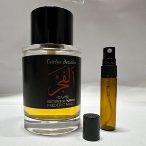 Puede incluir: Un frasco de perfume de vidrio transparente con una tapa negra y un pulverizador más pequeño de color ámbar. El frasco principal tiene una etiqueta negra con escritura árabe y el nombre "Carlos Benaïm". El perfume es de color dorado.
