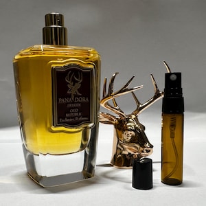 May include: A perfume bottle with a gold cap and a label that reads "Pana Dora Sweden Oud Republic Exclusive Perfume." A small, amber-colored spray bottle and a gold deer head figurine are also present.