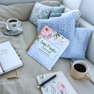 May include: A white and blue floral journal with the title "Iman Practice Journal (Islamic Journal)" on a light beige surface with a cup of coffee, a pen, and a notebook.