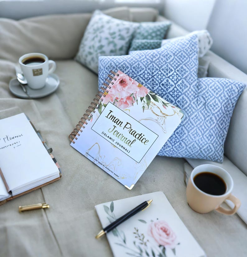 May include: A white and blue floral journal with the title "Iman Practice Journal (Islamic Journal)" on a light beige surface with a cup of coffee, a pen, and a notebook.