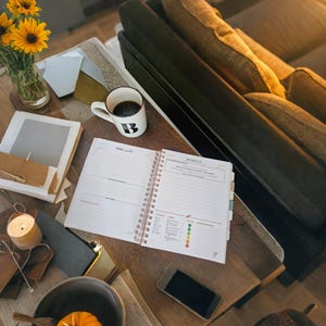 May include: A wooden table with a white planner, a cup of coffee, a smartphone, a candle, a bowl with a pumpkin, and a bouquet of yellow flowers. The table is next to a brown sofa.
