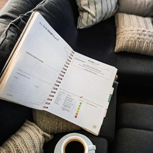 May include: A white spiral bound journal with a black and gold design. The journal is open to a page with a section for daily tasks and a section for mood tracking. A cup of coffee and a sugar cube are on a white saucer next to the journal.