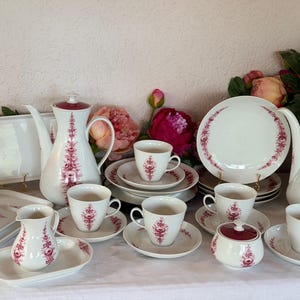 Op de afbeelding: Een theeservies van wit porselein met een delicaat magenta bloemmotief. De collectie bevat twee hoge theepotten, meerdere kopjes met bijpassende schoteltjes, een melkkan, een suikerpot en rechthoekige serveerbladen, allemaal samen opgesteld.