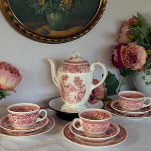 Könnte beinhalten: Ein cremefarbenes Porzellan-Teeservice mit einem roten Transferdruck-Muster aus ländlichen Landschaften und Blumenmotiven. Das Set besteht aus einer Teekanne mit Deckel, drei Teetassen und drei passenden Untertassen, alle mit roten Verzierungen.