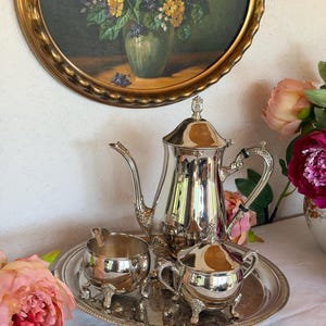 Puede incluir: Un juego de té plateado que consta de una tetera alta, un azucarero con tapa y una jarra de leche abierta, todo dispuesto sobre una bandeja ovalada de plata a juego. Cada pieza presenta asas curvas ornamentadas y pies decorativos.