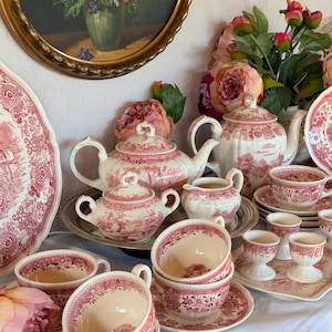 May include: A collection of cream-coloured ceramic tea service pieces featuring a red transferware pattern of pastoral scenes and floral borders, including teapots, a sugar bowl, a creamer, teacups with saucers, and egg cups on a white cloth.