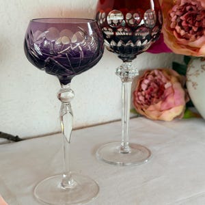 Può includere: Due calici da vino in cristallo con lunghi steli trasparenti e basi rotonde. Un calice è viola intenso con motivi floreali intagliati, mentre l'altro è rosso rubino profondo con un design geometrico a cerchi intagliato sulla coppa.