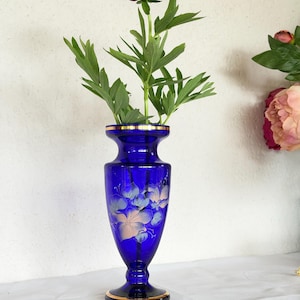May include: A cobalt blue glass vase with a pedestal base and a flared rim, featuring a gold-painted trim and a delicate white floral design on the body. A single dark red peony with green leaves is placed inside the vase.