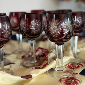 Puede incluir: Una colección de copas de vino de cristal tallado en color rojo intenso con intrincados patrones de estrellas grabados en los cuencos. Cada copa tiene un tallo de cristal transparente y una base circular, destacando el contraste entre el vidrio rubí y el diseño.