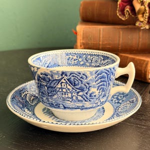 Puede incluir: Un juego de taza y platillo azul y blanco. La taza y el platillo presentan un diseño de paisaje detallado. La taza tiene un asa y se asienta sobre un platillo a juego. El juego es de cerámica y de estilo vintage.
