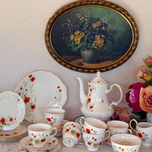 Könnte beinhalten: Ein Teeservice aus weißem Porzellan mit einem zarten Muster aus roten Mohnblumen und grünem Blattwerk sowie Goldrand, bestehend aus einer Teekanne, mehreren Tassen, Untertassen und Tellern, arrangiert neben einem großen ovalen Blumengemälde.