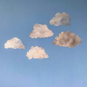Könnte beinhalten: Fünf einzelne wolkenförmige Wandaufkleber mit einer strukturierten, metallisch silbernen und grauen Oberfläche. Jede Wolke hat einen unregelmäßigen, gewellten Rand und ein Pinselstrichmuster, angeordnet in einer lockeren Formation auf blauem Grund.