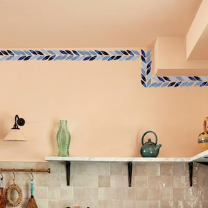Könnte beinhalten: Küchenszene mit einer pfirsichfarbenen Wand, verziert mit einem blauen Blättermuster. Ein weißes Regal beherbergt eine grüne Glasflasche, eine türkisfarbene Teekanne und eine dekorative Vase. Eine Wandlampe und Küchenutensilien ergänzen die Einrichtung.