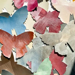 Könnte beinhalten: Eine Sammlung farbenfroher Schmetterlingsausschnitte in verschiedenen Blau-, Rot-, Grau- und Grüntönen. Die Schmetterlinge haben ein strukturiertes, verwittertes Aussehen und sind in einem geschichteten Muster angeordnet. Der Hintergrund ist weiß.